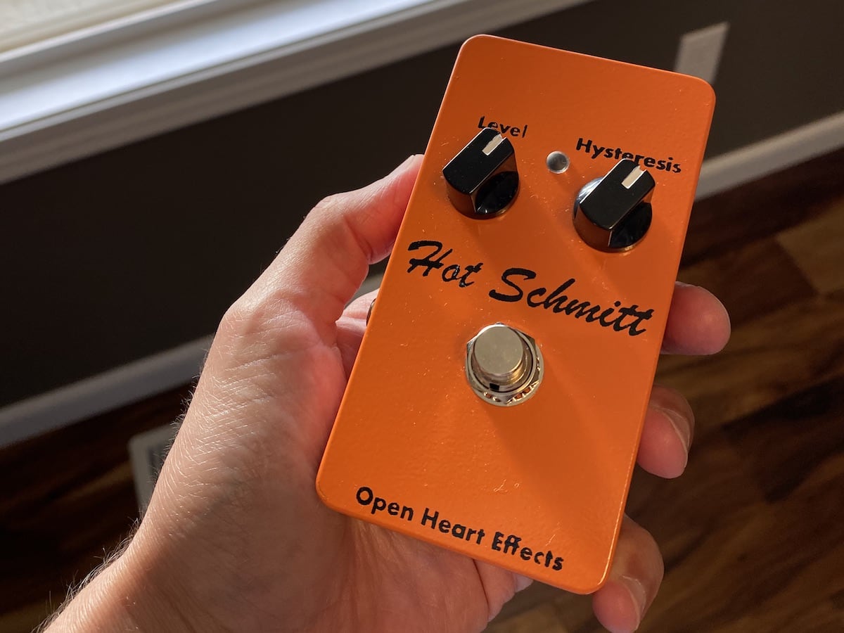 A hand holds a guitar
pedal built in an orange Hammond 1590B enclosure. The pedal has two knobs
labeled 'Level' and 'Hysteresis'. There are also a small light and a large
chrome stomp switch on the front of the enclosure. Right in the middle, the
pedal is labeled 'Hot Schmitt' in a swaggering brush script. At the bottom, it
says 'Open Heart Effects' in a modern sans-serif typeface.