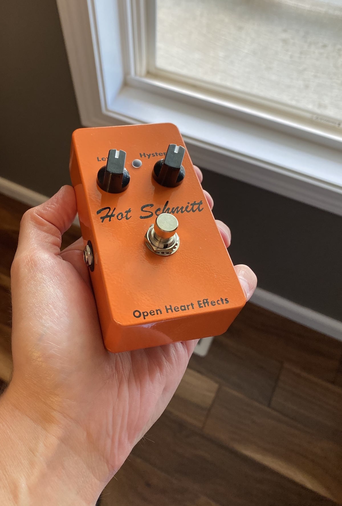 A hand holds a vivid orange
guitar effect pedal up to the evening summer light streaming in a west-facing
window. The deep black knobs and chrome footswitch gleam in the fading light.
Black silkscreened markings declare that this is the Hot Schmitt from
Open Heart Effects. The knobs are labeled Level and Hysteresis.